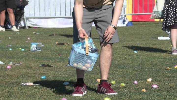 lower half of a body of a person bending over picking up an egg in a park while holding a basket filled with eggs